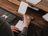 Person taking notes or tasks at a desk with a laptop and keyboard |-