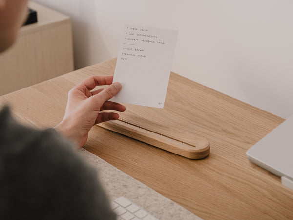 Person holding a paper to-do list at a wooden desk with a laptop in the background |-