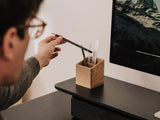 Person reaching for a pencil from a wooden pen holder on a desk in front of a computer screen. | oak